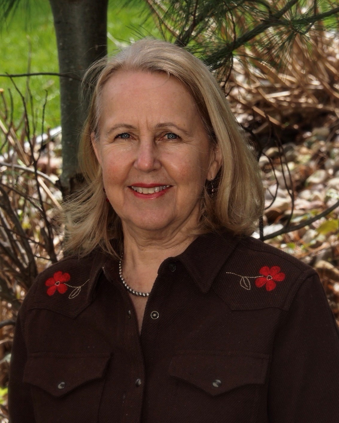 Portrait of Christine Skelly outdoors, standing in front of trees and foliage. Christine has shoulder-length hair and is wearing a dark brown button-up shirt with red floral embroidery. 